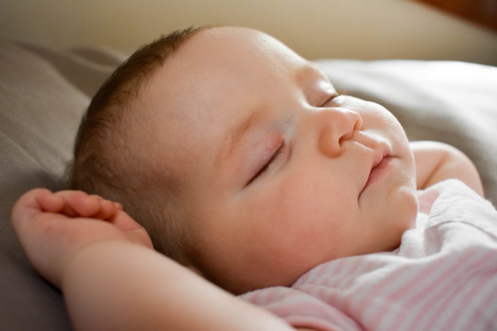 A baby is sleeping with their arms raised next to their head. The baby's hair is dark and they are wearing a pink and white short sleeve shirt.